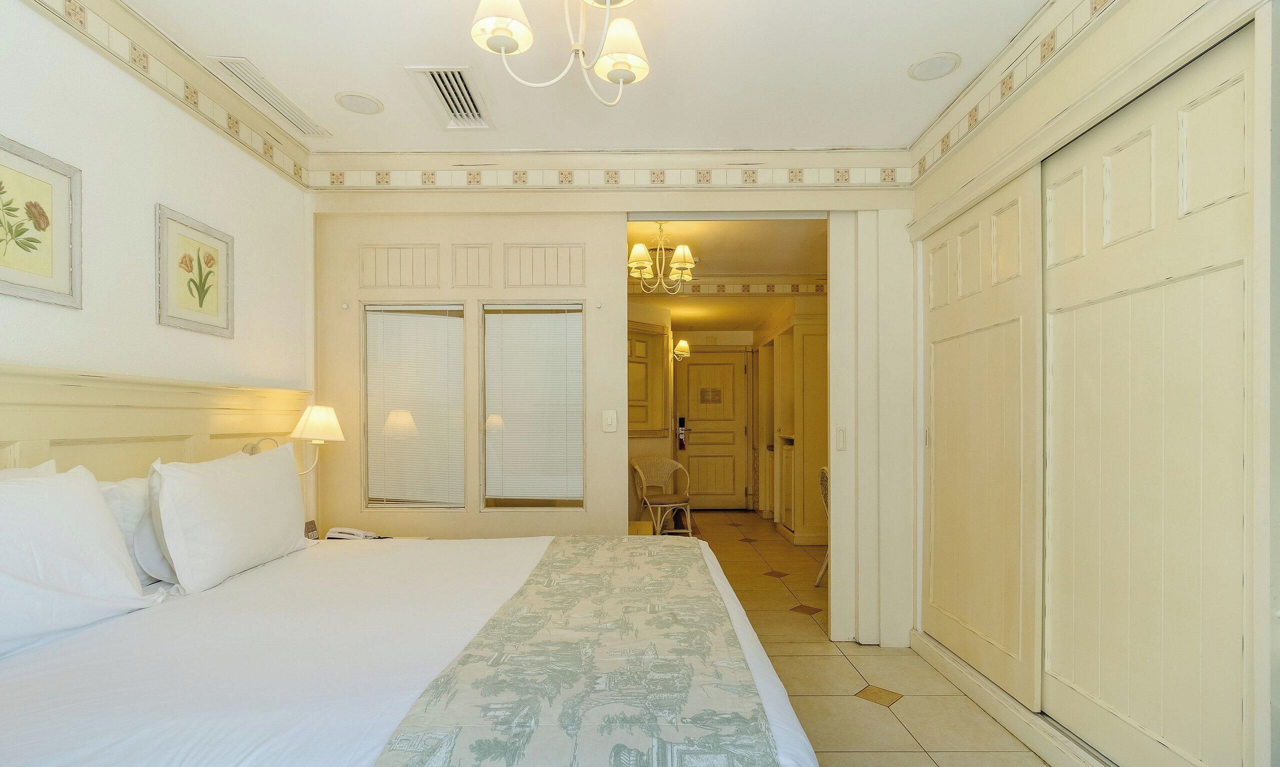 A hotel room with a bed, bedside lamps, a window with blinds, a door leading to a hallway, and a chandelier.