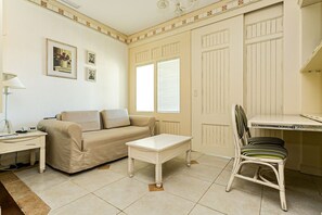 A room with a beige sofa, a small white table, a wicker chair, and a desk with a chair. There are pictures on the wall and a radiator on the wall. - Charmosos Studios no IL Campanario Villaggio Resort ILCJR II (Florianópolis)