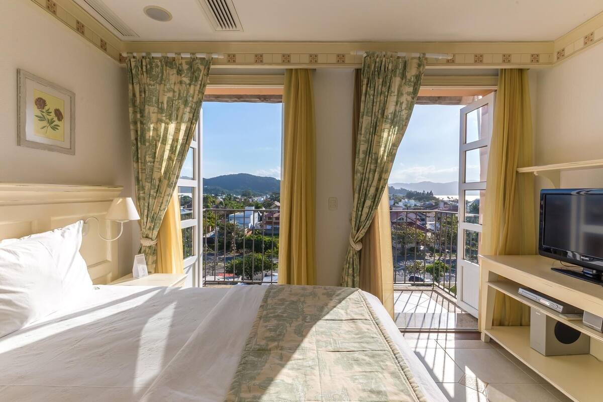 Una habitación de hotel con cama, televisión y balcón con vistas a la ciudad y montañas.