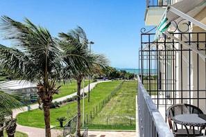 A balcony with a view of a grassy pathway and palm trees, leading to a building with a gate.