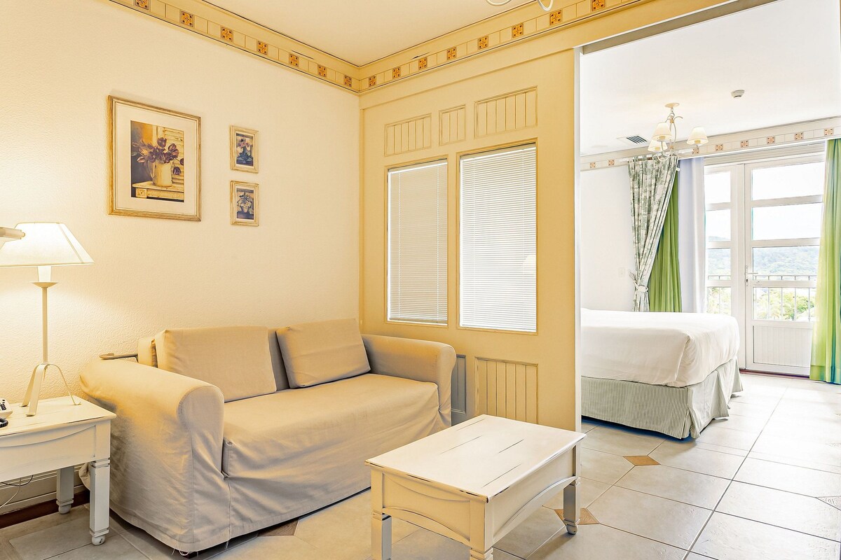 Quarto com sofá, uma mesinha e uma cama. Janelas com persianas oferecem vista para o exterior.