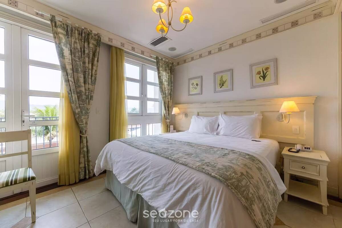 Un dormitorio con cama, mesitas de noche, una silla y una ventana con cortinas.