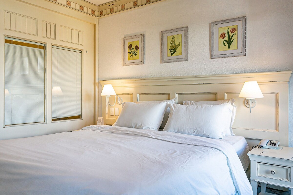 Cama bem arrumada com lençóis brancos, duas luminárias de cabeceira e mesa de cabeceira com telefone.