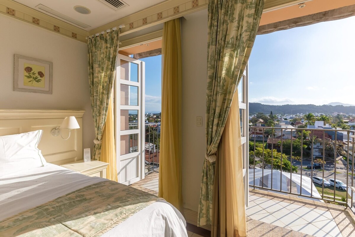 Quarto de hotel com cama, mesa de cabeceira, janela com cortinas e vista para a cidade.