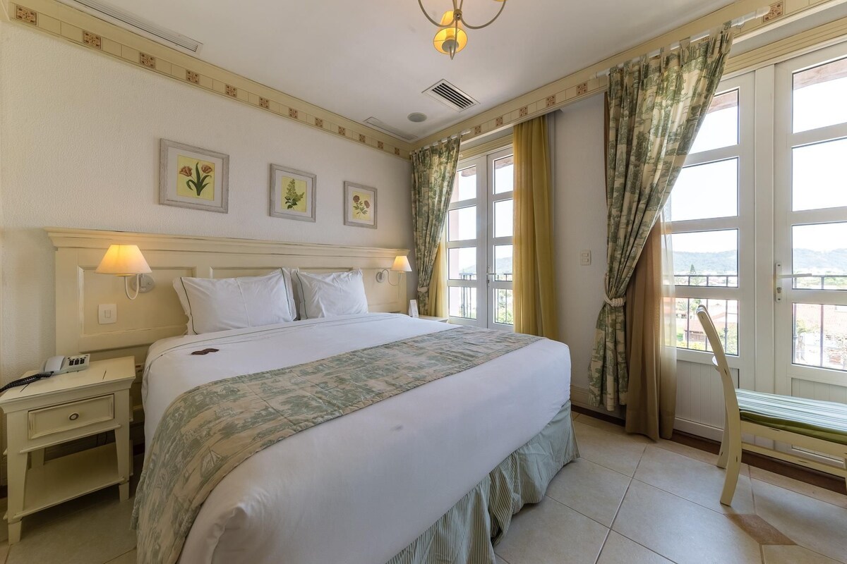 Quarto de hotel bem arrumado com uma cama grande, mesas de cabeceira, escrivaninha e uma cadeira. Há cortinas nas janelas e obras de arte nas paredes.