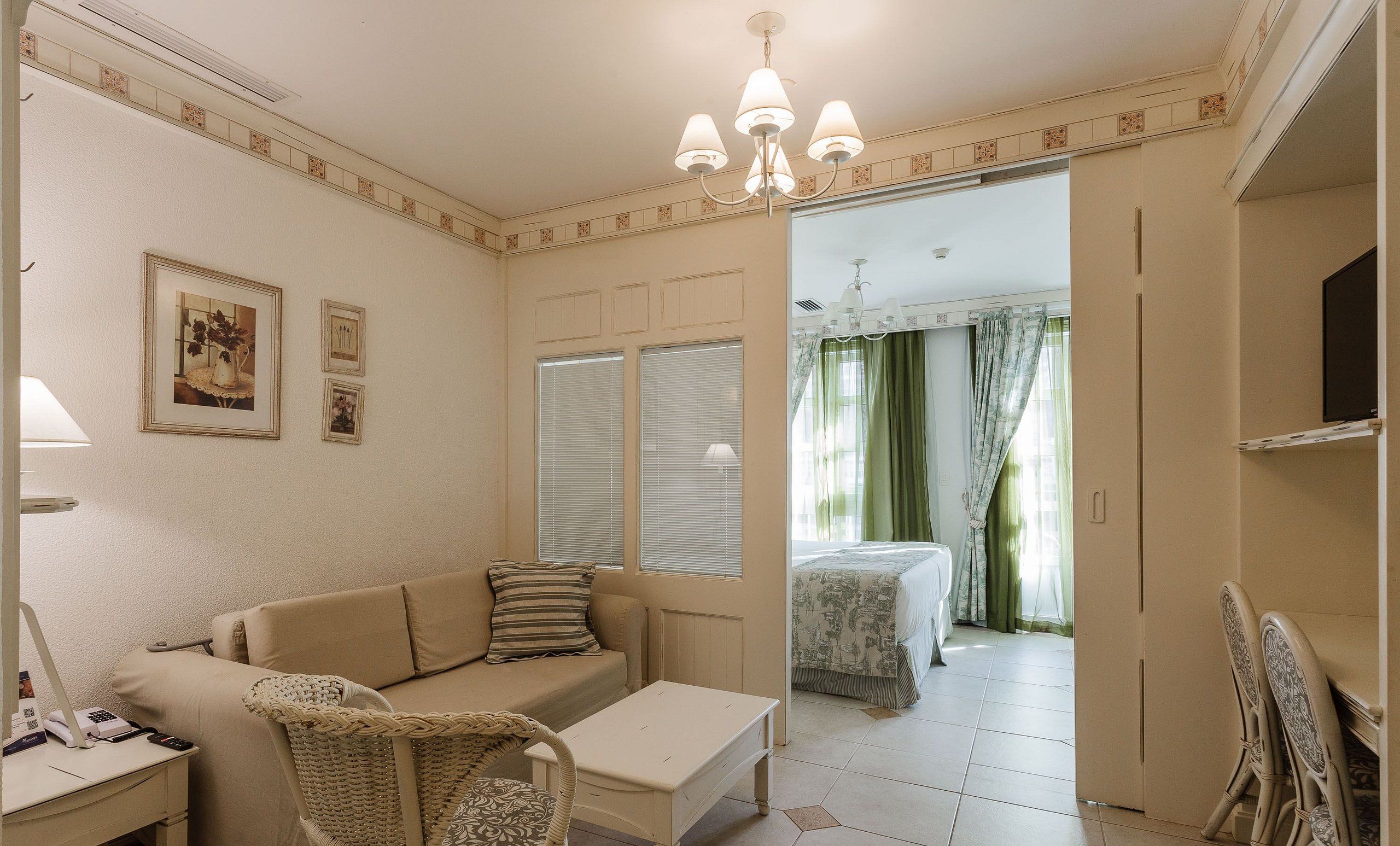 A room with a sofa, a small table, a wicker chair, a bed with a patterned bedspread, and a chandelier.