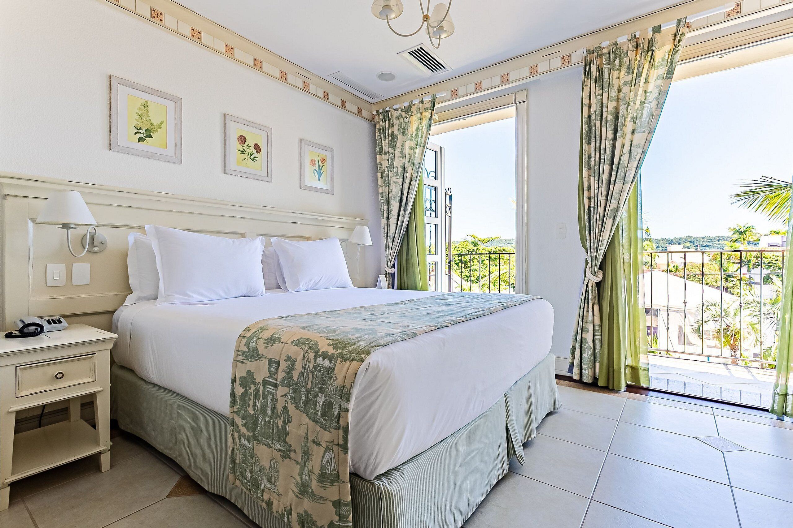 A hotel room with a large bed, a nightstand, a balcony with a view of the city, and two wall-mounted lamps.