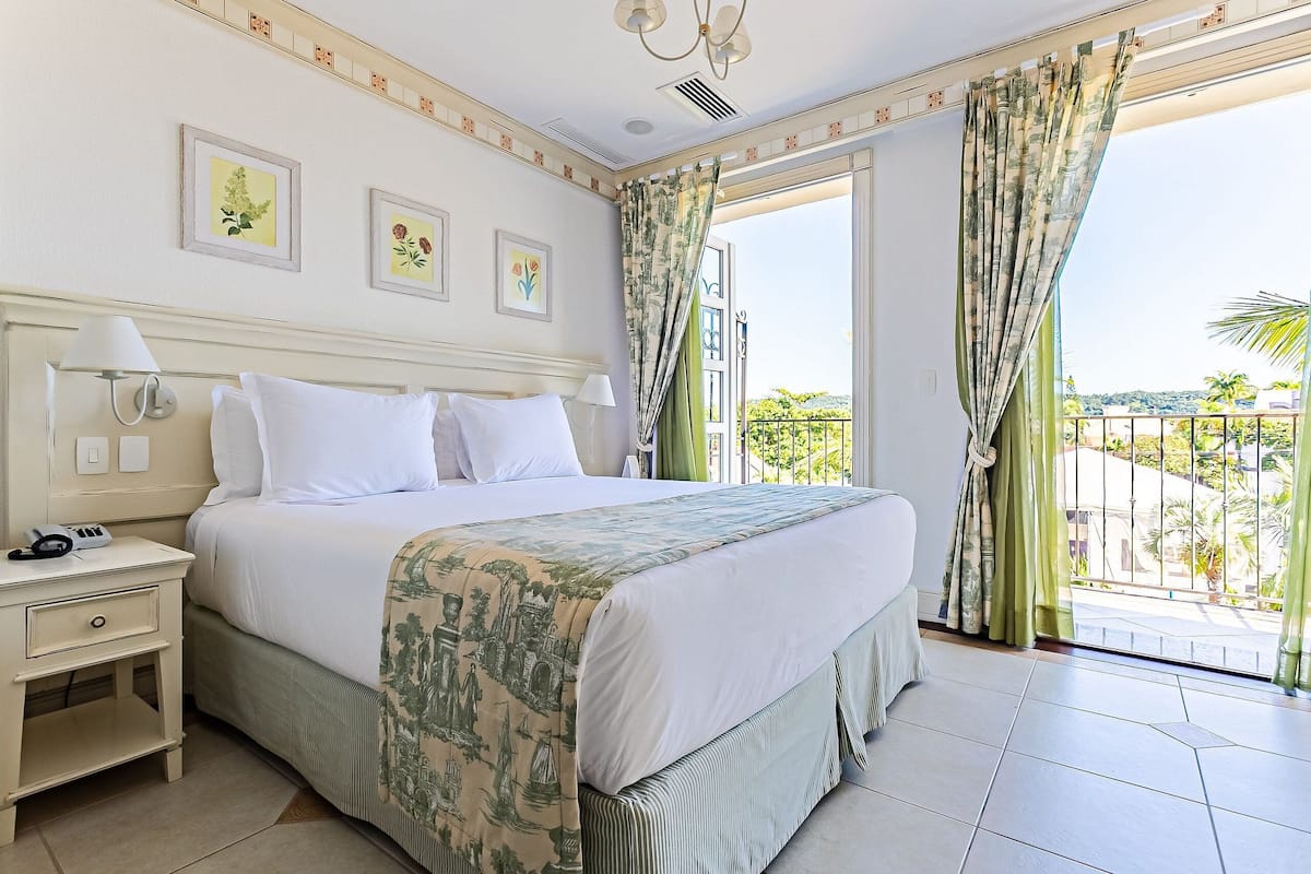 A hotel room with a large bed, a nightstand, a balcony with a view of the city, and two wall-mounted lamps.