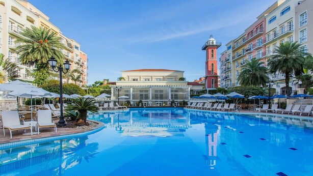 A swimming pool with lounge chairs and umbrellas, surrounded by multi-story buildings with balconies. - Charmosos Studios no IL Campanario Villaggio Resort ILCJR II (Florianópolis)