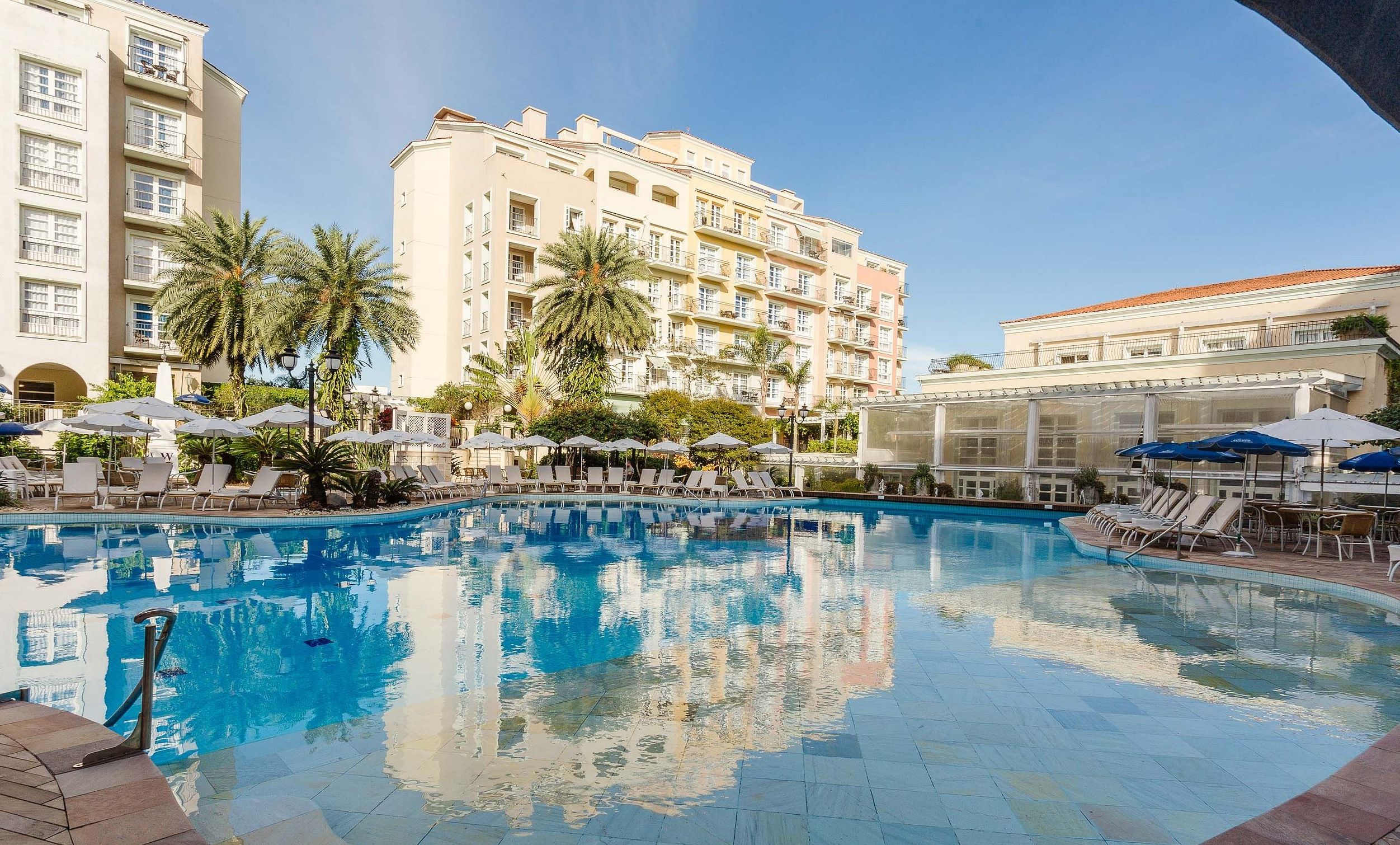 A swimming pool with lounge chairs and umbrellas, surrounded by palm trees and hotel buildings.