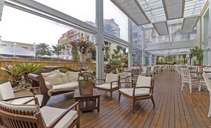 Um pátio interno amplo, com piso de madeira, assentos acolchoados brancos e grandes janelas com vista para os edifícios ao redor.