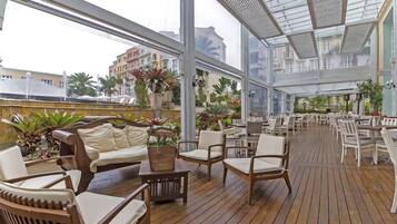 Un patio interior amplio con suelo de madera, asientos acolchados blancos y grandes ventanales que ofrecen vistas de los edificios circundantes.