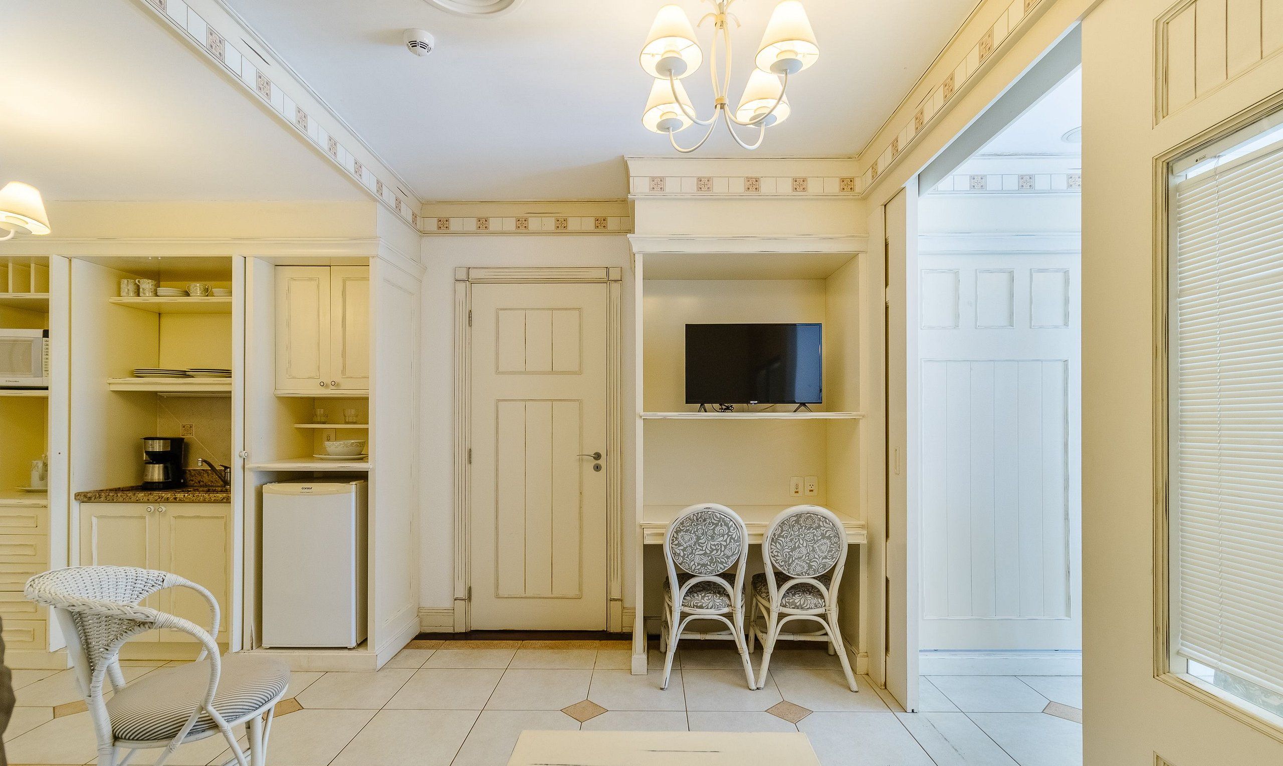 Eine kompakte Küche mit weißen Schränken, einer Mikrowelle, einer Kaffeemaschine und einem Kühlschrank. Zwei Korbstühle und ein kleiner Tisch bieten Platz. Ein Fernseher ist über einem Regal angebracht.
