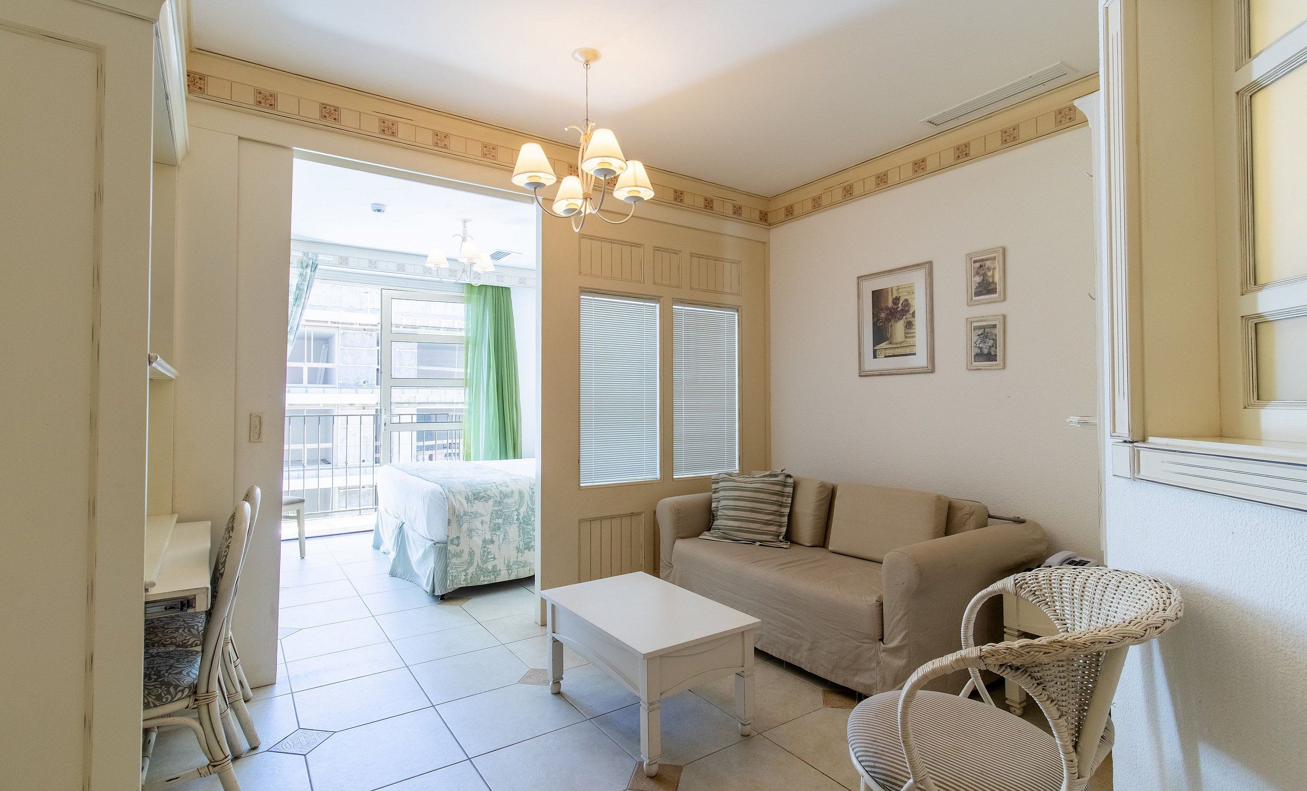 Una habitación con sofá, una mesita, una silla de mimbre, ventana con persianas y un candelabro.