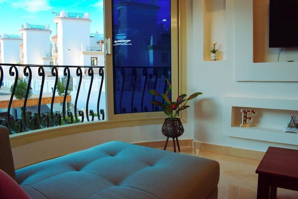 Uma sala de estar com uma janela grande, um sofá, uma mesa pequena e uma TV fixada na parede.