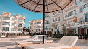 Et poolområde med liggestole og en stor parasol, omgivet af høje bygninger med altaner.