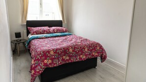 A bedroom with a bed, a bedside table, a lamp, and a window with curtains. - Victoria Road Retreat - Charming 2 Bedroom House (Barnsley)