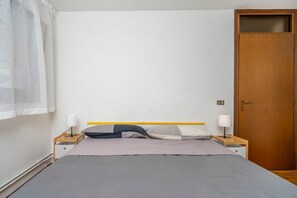 Ein Schlafzimmer mit einem Bett, zwei Nachttischen mit Lampen, einer Holztür und einem Fenster mit Vorhängen.
