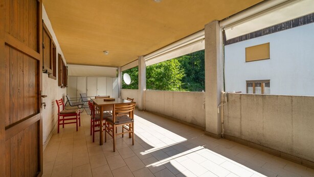 A covered outdoor area with a dining table and chairs, a wooden door, and a view of greenery through the windows. - GuestHost - Cozy Apartment with Terrace - Tarvisio (Tarvisio)