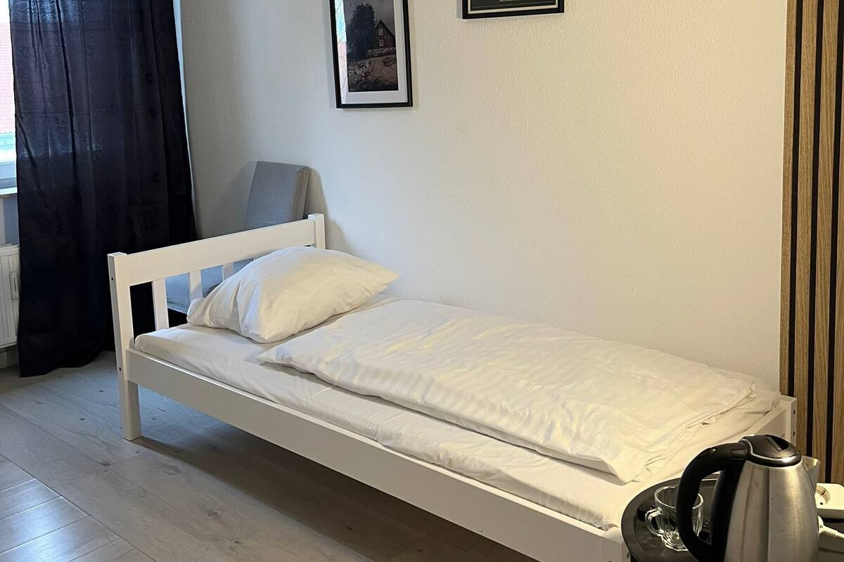 Ein Zimmer mit Einzelbett, einem kleinen Tisch mit Wasserkocher, einem gerahmten Bild und einem Fenster mit Vorhängen.