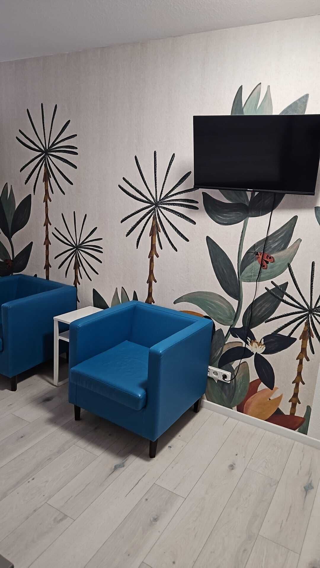 Une pièce comprenant deux fauteuils bleus, une table d’appoint blanche, un téléviseur à écran plat fixé au mur et une fresque représentant un feuillage tropical.