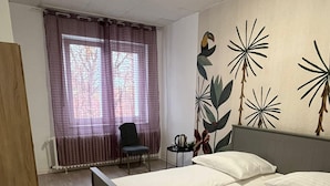 Une chambre d’hôtel avec un lit, une chaise, une petite table avec une bouilloire et une fenêtre avec des rideaux.