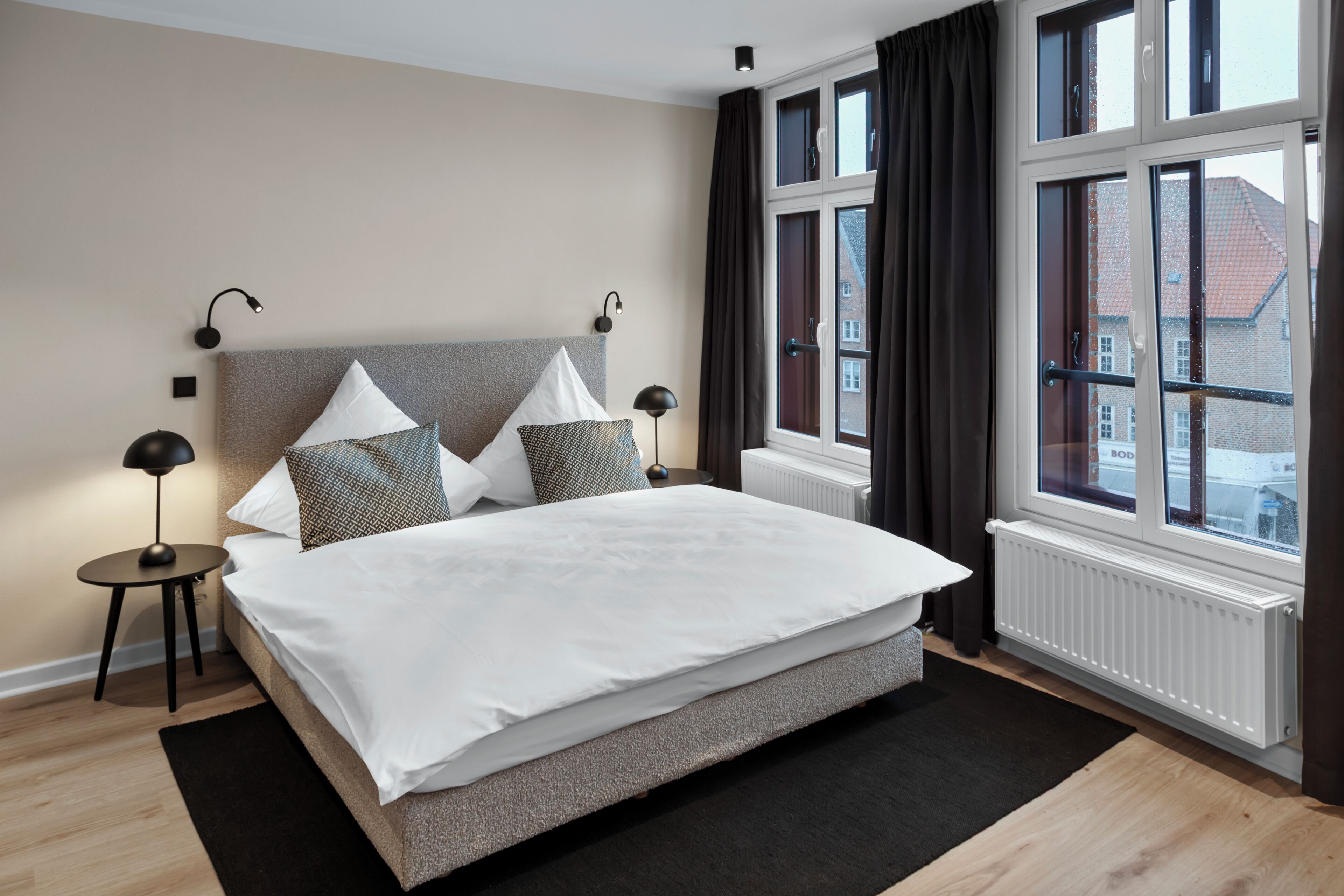 A modern bedroom with a large bed, two bedside lamps, a radiator, and a window with curtains.