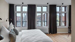 Ein modernes Schlafzimmer mit einem großen Bett, zahlreichen Fenstern und Blick auf umliegende Gebäude.