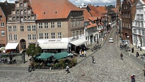 Eine Kopfsteinpflasterstraße gesäumt von historischen Gebäuden, ein zentraler Platz mit einer grünen Markise und ein Kirchturm in der Ferne.
