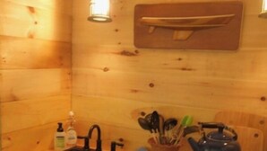 Interior de una cabaña de madera con zona de cocina, que incluye un fregadero, una tetera sobre la estufa y un estante con utensilios de cocina.