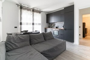 A modern living room with a grey sofa, a dining table, and a kitchen area with dark cabinets and a built-in oven. - GuestHost - Cozy 50m2 apartment with balcony, located on the seventh floor of a building with a lift, in a residential context surrounded by greenery.The area is very quiet and well connected thanks to local public transport: a 10-minute walk away is the (Milano)