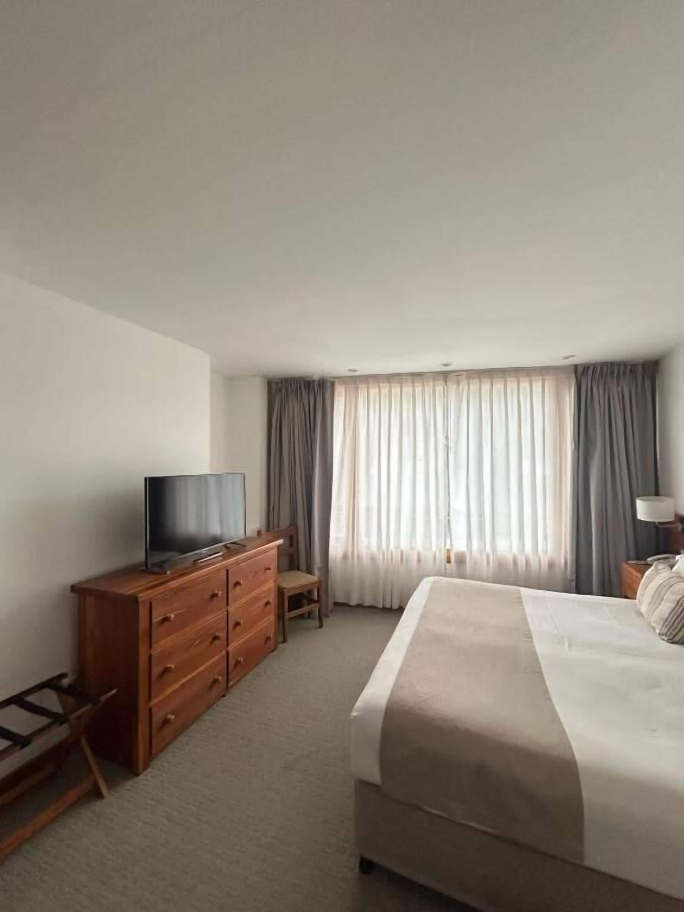 A hotel room with a bed, a wooden dresser, a television, and a window with curtains.