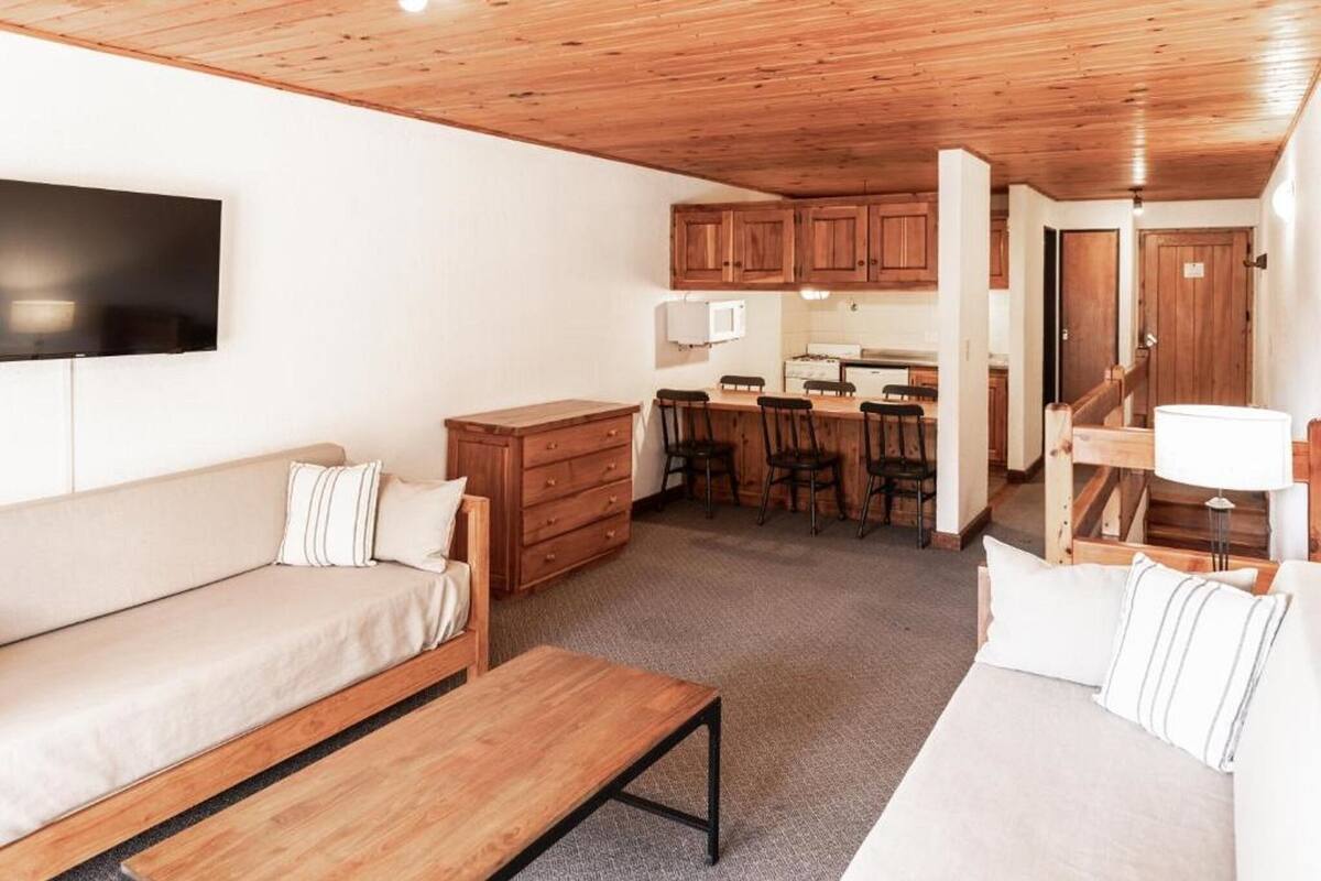 A cozy living room with a sofa, a wooden bench, and a kitchen area with wooden cabinets and a dining table.