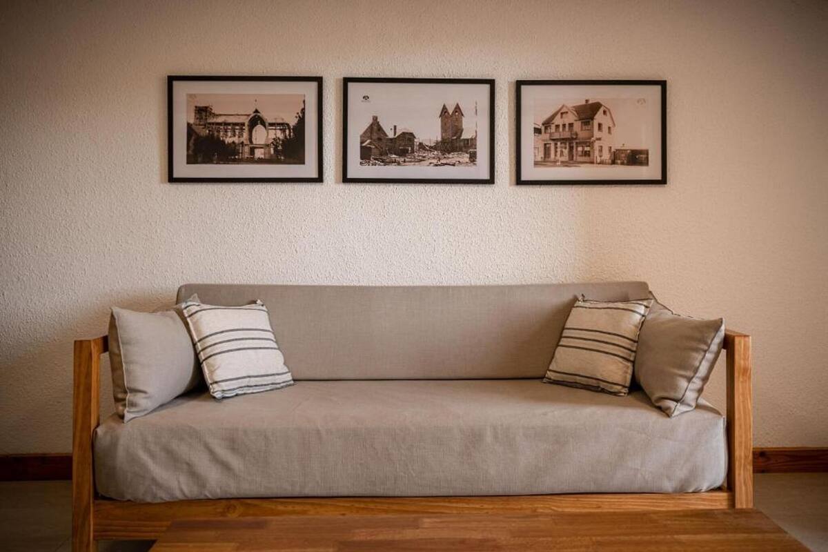 A wooden sofa with a beige cushion, flanked by two pillows with striped patterns, against a plain wall. Three framed black-and-white photographs are hung above the sofa, depicting architectural structures.