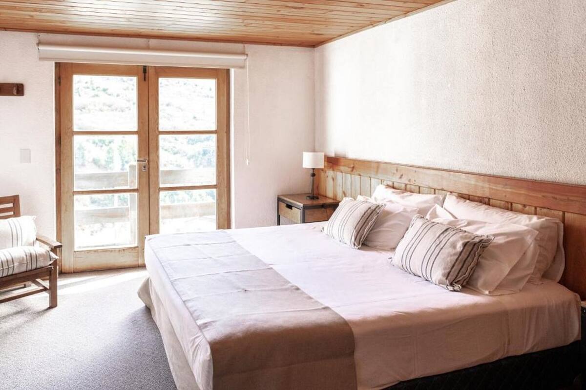 A bedroom with a large bed, a wooden headboard, a nightstand with a lamp, and a window with a view of greenery.