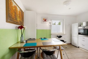 Una habitación pequeña y luminosa con una mesa de madera, sillas, un jarrón con flores, un cuadro y una kitchenette.
