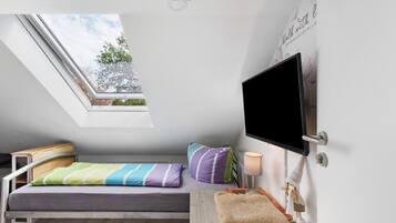 Une petite chambre avec un lit simple, une lucarne, un téléviseur fixé au mur et une table de chevet avec une lampe.