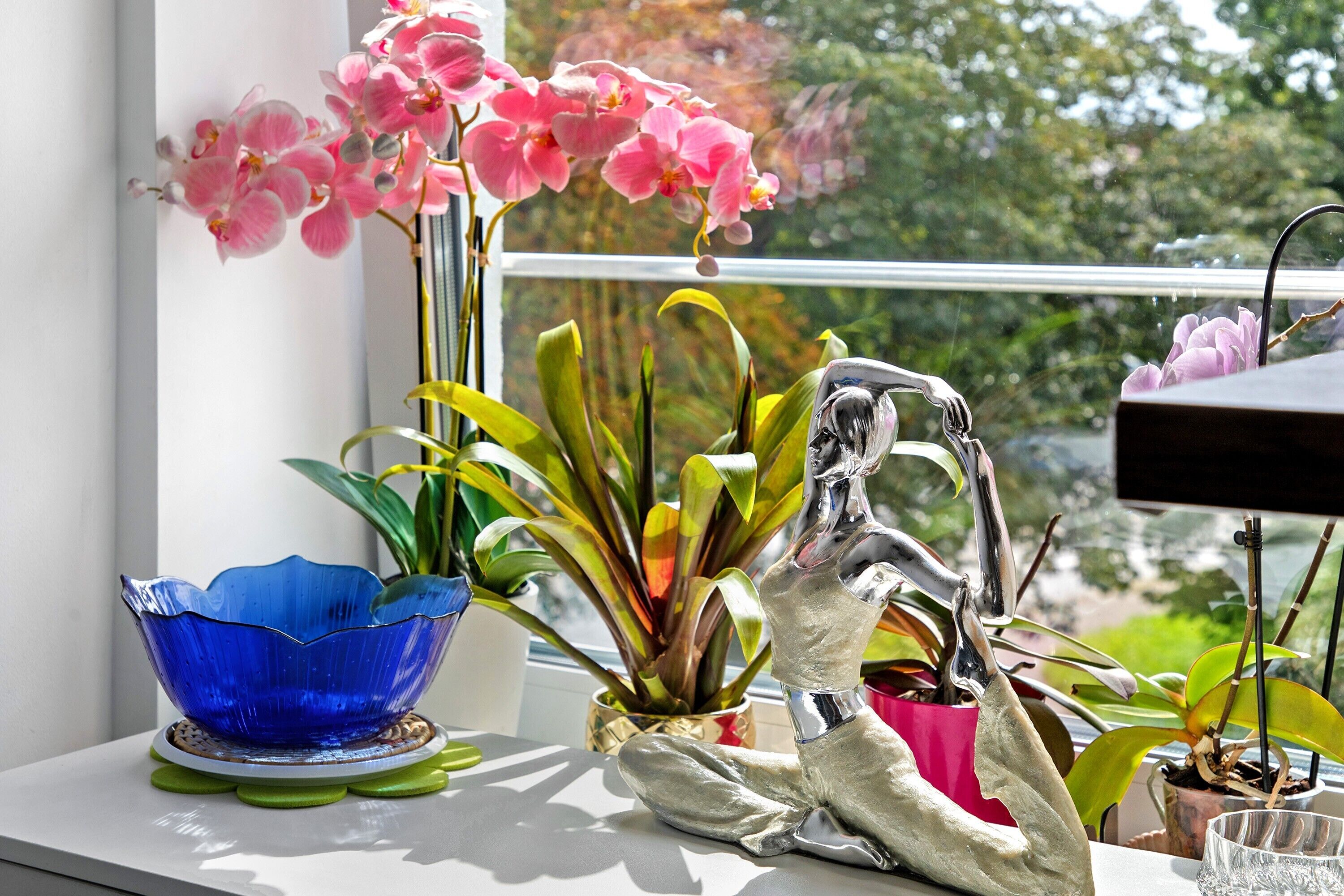 A window sill with a blue glass bowl, a silver sculpture, and pink orchids.