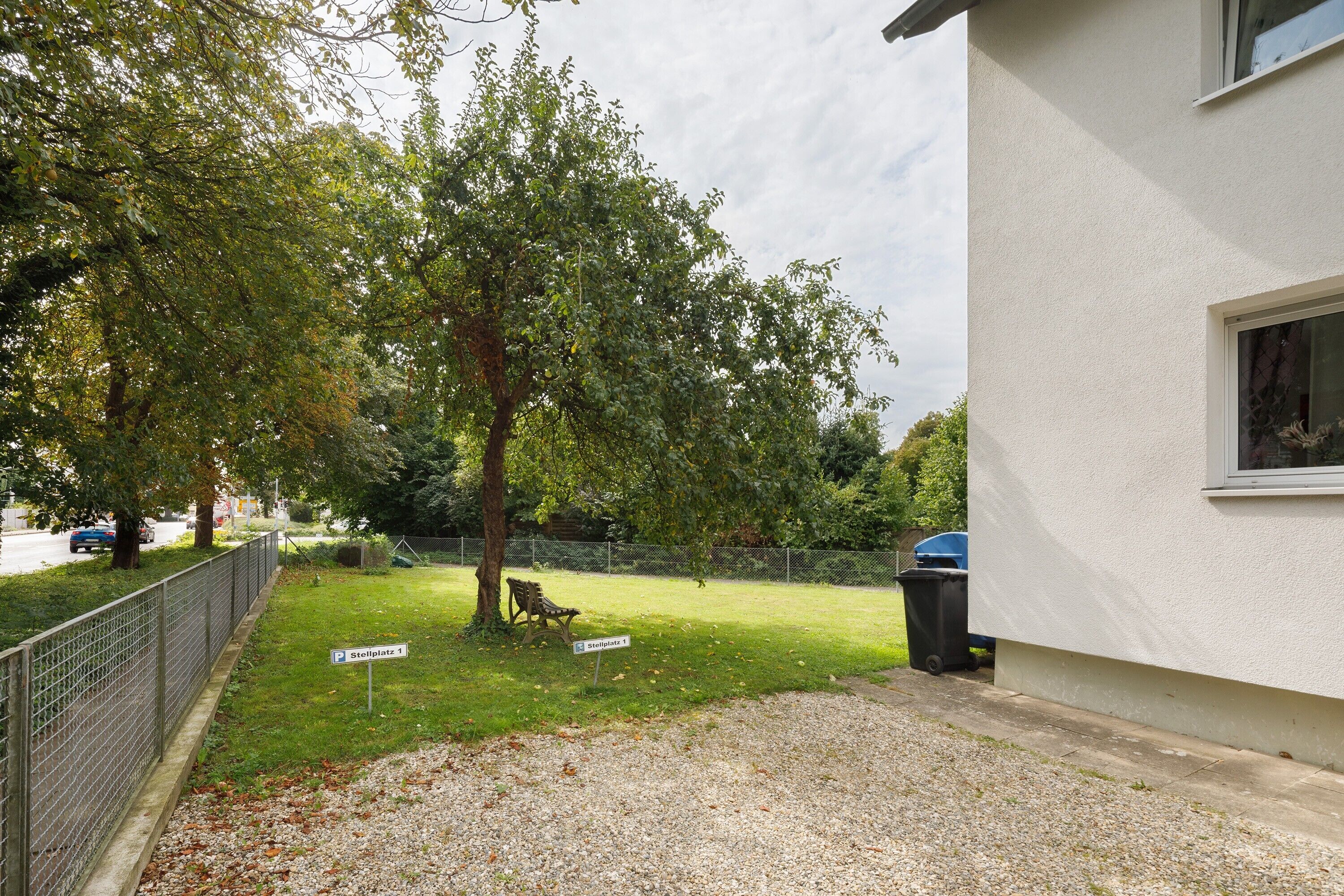 A residential area with a paved driveway, a bench, a tree, and a building with a window.