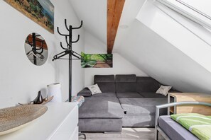 Un dormitorio estilo loft con un sofá seccional gris, una mesita de noche pequeña, techo con vigas de madera y un cuadro de un paisaje forestal.