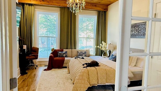 A bedroom with a wooden ceiling, a chandelier, a bed with a beige quilt, a red chair, and a large window with curtains.