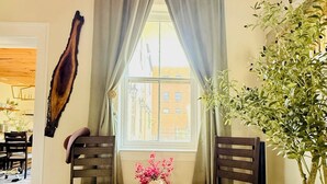 A room with wooden flooring, two dark wooden chairs, a small round table with a vase, a large window with curtains, and a wall-mounted plant.