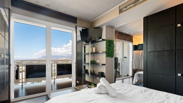 Een moderne slaapkamer met een groot bed, een balkon met uitzicht op de stad, inbouwplanken en een donkere garderobe.