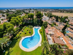 A swimming pool with a curved design, surrounded by greenery and palm trees, with a view of the sea and residential buildings in the background. - Apartment 'Con Vistas Al Mar' with Sea View, Wi-Fi and Air Conditioning (Marbella)