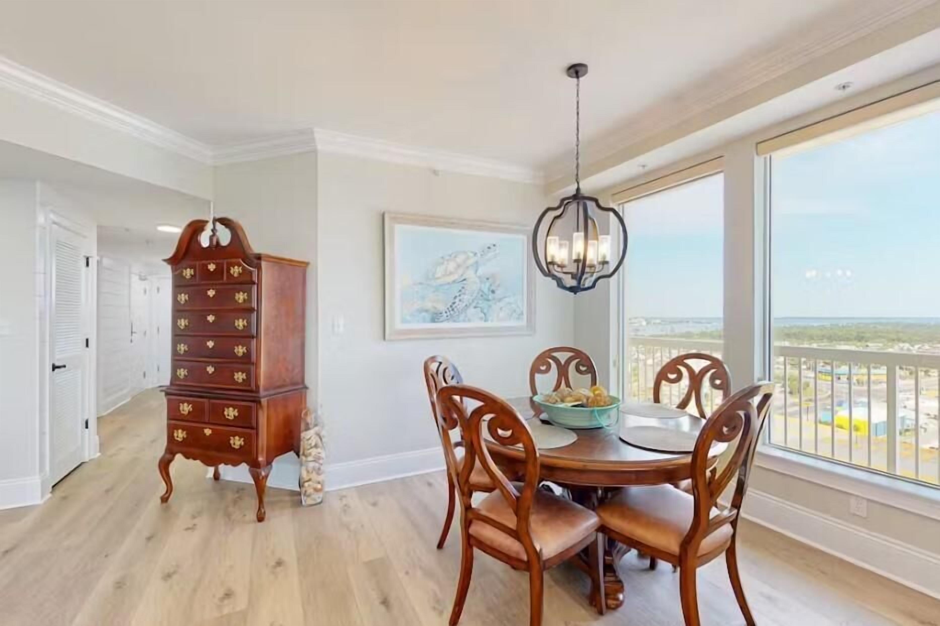 A dining area with a wooden table and chairs, a chandelier, and a view of the ocean through large windows.