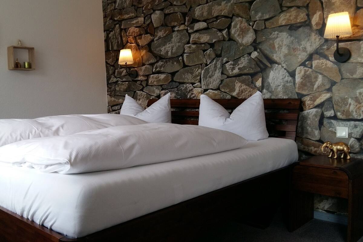Un lit avec du linge de lit blanc, dans une pièce dont un mur est en pierre et qui possède une tête de lit en bois.
