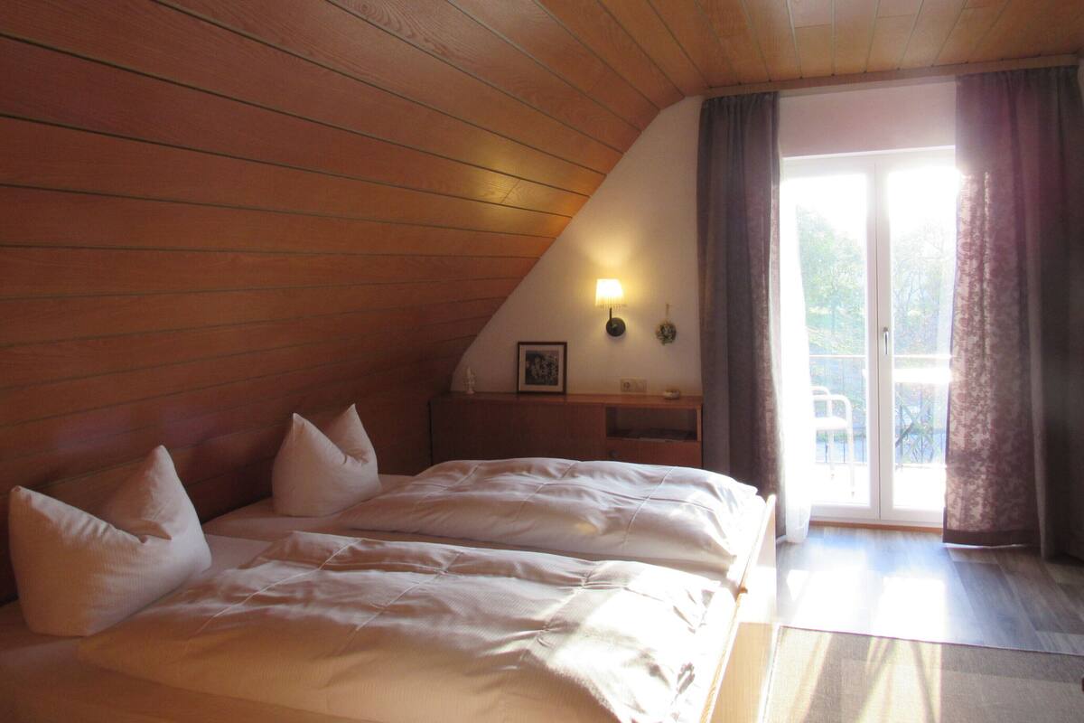 Une chambre avec un plafond en bois, un lit recouvert de linge de lit blanc, une table de chevet avec une lampe, et une vue à travers une porte vitrée.