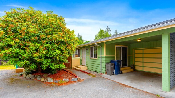 Une maison de plain-pied à l’extérieur vert, avec un garage et un petit jardin devant, où se trouve un arbre.