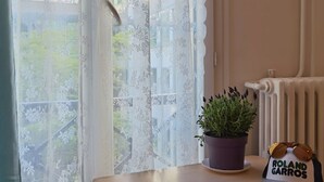 Una habitación con una ventana de encaje, un radiador, una planta en maceta y un gorro con texto.