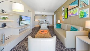 A modern hotel room with a flat-screen TV, a wooden coffee table, a sofa, and a kitchen area in the background.